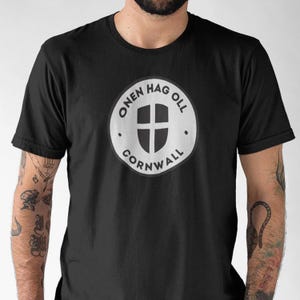 May include: Black t-shirt with a white circular emblem. The emblem has the text "ONEN HAG OLL CORNWALL" and a shield with a cross. The shirt is a crew neck style, a common design in the UK.
