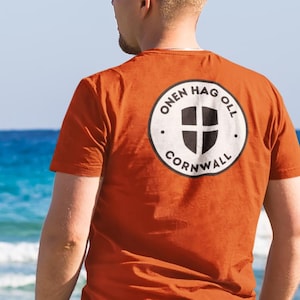 Può includere: T-shirt arancione bruciato con grafica circolare sul retro. La grafica presenta uno scudo nero con una croce bianca e le parole "ONEN HAG OLL CORNWALL" in un motivo circolare. La maglietta è a maniche corte.