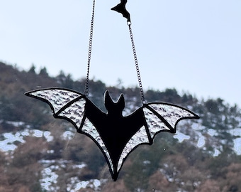 May include: A black bat-shaped suncatcher hanging from a chain. The bat has a detailed wing design with a textured, iridescent appearance. A smaller bat silhouette hangs above. The background shows a blurred outdoor scene with trees and a snowy landscape.