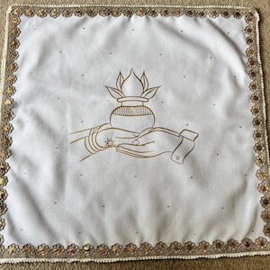 May include: White square cloth with a gold embroidered border. The centre features a gold illustration of hands holding a pot with a floral design. Small, scattered rhinestones add sparkle.