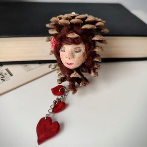 Könnte beinhalten: Ein skurriles handgefertigtes Ornament mit einem Kiefernzapfenkopf, bemaltem Gesicht, roten Lippen und einer rosa Blume. Das Ornament hat eine Kette aus drei roten Herzanhängern. Der Kiefernzapfenkopf hat braunes Haar.