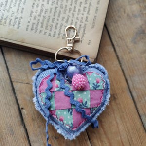 以下が含まれることがあります： パッチワークデザインのハート型キーホルダー。デニムの縁取り、ピンクとグリーンの花柄生地、ビーズの装飾が施されています。上部には金色の留め金が付いています。