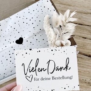 Könnte beinhalten: Eine Geschenkbox mit weißem Seidenpapier mit schwarzen Punkten. In der Box befinden sich Trockenblumen und eine Karte mit dem Schriftzug "Vielen Dank für deine Bestellung!" in schwarzer Schrift und einem Herz.