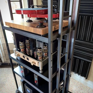 Puede incluir: Un rack de equipos de audio de varios niveles con un tocadiscos rojo en la parte superior. El rack tiene un marco de metal negro y estantes de madera. Se ven varios componentes de audio.