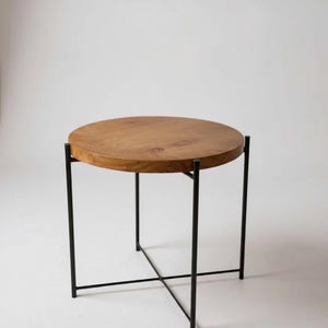 Könnte beinhalten: Ein runder Beistelltisch mit einer Tischplatte aus Naturholz und schwarzen Metallbeinen. Der Tisch hat ein schlichtes, modernes Design, wobei die Beine an der Basis ein X bilden, um Stabilität zu gewährleisten. Die Holzplatte hat einen Durchmesser von ca. 50 cm.