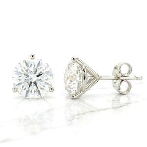 May include: A pair of stud earrings featuring round, brilliant-cut clear stones set in polished silver-toned metal. One earring is shown from the front, displaying the faceted stone, while the other is shown from the side, revealing the three-prong setting.