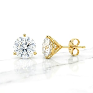 May include: A pair of stud earrings featuring round, brilliant-cut clear stones set in yellow gold. One earring is shown from the front, displaying the faceted stone, while the other is shown from the side, revealing the gold basket setting.
