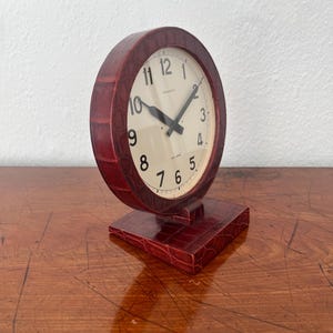 Puede incluir: Un reloj analógico redondo de color granate con una esfera color crema y números negros. El reloj tiene un patrón texturizado, similar a la piel de cocodrilo, en su carcasa y base. El reloj está sobre una superficie de madera.