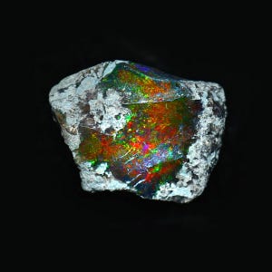 May include: A raw, unpolished opal gemstone featuring a dark, textured matrix with a vibrant play-of-color. The stone displays intense flashes of red, orange, green, and blue light across its fractured, crystalline surface against a dark background.