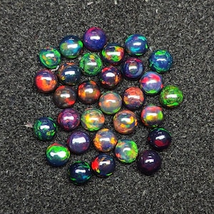 以下が含まれることがあります： 緑、青、赤、黒の様々な色合いの虹色のオパールカボションが25個集められています。カボションは暗い灰色の表面に円形に配置されています。