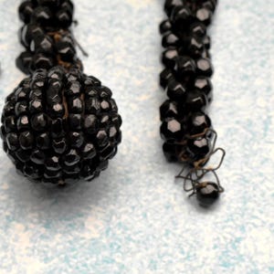 May include: A close-up of a black beaded necklace with a round, black bead at the end. The beads are small and tightly packed together, creating a smooth, uniform surface.
