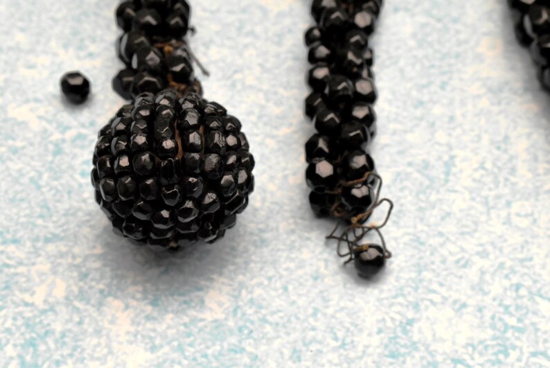 May include: A close-up of a black beaded necklace with a round, black bead at the end. The beads are small and tightly packed together, creating a smooth, uniform surface.