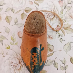 May include: A wooden shoe-shaped pincushion with a brown leather exterior and a soft, tan cushion. The shoe features a painted windmill scene in teal and gold, with a jute string attached for hanging.