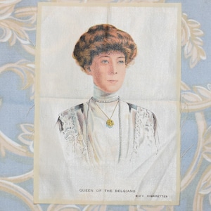 Könnte beinhalten: Eine Vintage-Zigarettenkarte mit einem Porträt von Königin Elisabeth von Belgien. Die Karte ist weiß mit einem goldenen Rand und dem Text "Queen of the Belgians B.D.V. Cigarettes".