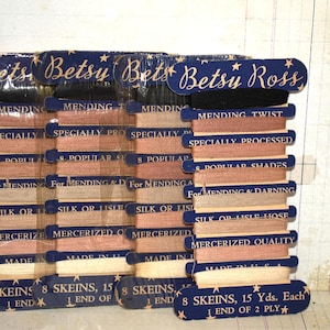 Puede incluir: Un conjunto de carretes de hilo de coser Betsy Ross. Los carretes son de varios tonos de marrón, beige y negro. Cada carrete está etiquetado con texto como "Mending Twist" y "Mercerized Quality".