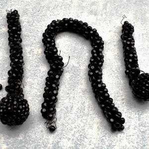 May include: Three black beaded necklaces with a round bead at the end of each. The necklaces are made of small, black beads that are strung together in a tight, spiral pattern.