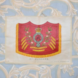 Könnte beinhalten: Eine Vintage-Zigarettenkarte aus Seide mit dem Wappen der 15. Husaren. Das Wappen ist ein roter Schild mit einer goldenen Krone, einem goldenen Buchstaben "R" und der Zahl "15" in einem Kreis. Der Schild ist von einem goldenen Rand und einer goldenen Franse umgeben. Der Text "15th Hussars" ist unter dem Wappen gedruckt.