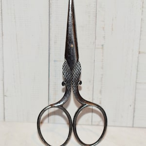 May include: Close-up of antique steel scissors. The scissors have a pointed tip and ornate handles with two finger holes. The metal has a dark, aged appearance, suggesting they are vintage. The blades are closed, and the design is symmetrical.