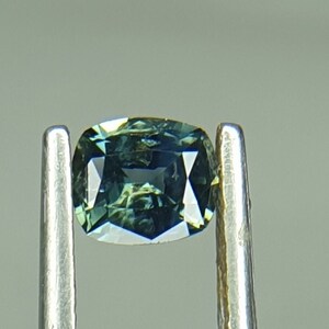 May include: A green and blue gemstone with a cushion cut. The gemstone is held in place by a pair of tweezers.