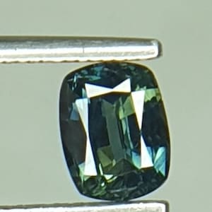 May include: A dark green gemstone with a cushion cut. The gemstone is held in place by a pair of tweezers.