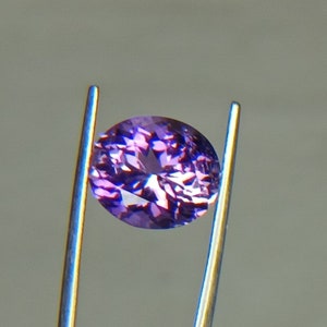 May include: A faceted, oval-shaped gemstone held by silver tweezers. The gem is a deep purple color with internal facets that reflect light. The tweezers are thin and metallic, contrasting with the gem's color. The background is a soft gray.