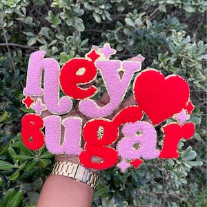 May include: Pink and red felt letters spelling "hey sugar" with a red felt heart and gold glitter accents.