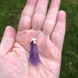 Può includere: Un ciondolo in cristallo di ametista viola con una ghiera in argento. Il ciondolo ha la forma di una punta e una superficie liscia e lucida.