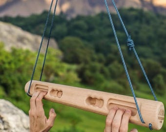 Puede incluir: Un fingerboard de madera con cuatro agarres de diferentes tamaños, colgando de una cuerda azul. El fingerboard está suspendido frente a una cordillera y árboles verdes.
