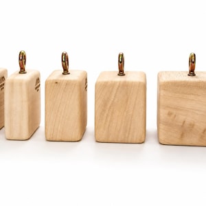 Peut inclure: Cinq blocs en bois clair d'épaisseurs variables disposés en rangée, chacun doté d'un boulon à œil en métal vissé au centre du sommet. Les blocs présentent une finition en grain de bois naturel lisse et un petit logo imprimé sur le côté.
