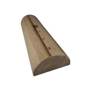 Puede incluir: Una presa de escalada de madera de color claro con una parte superior redondeada y una parte inferior plana. La presa tiene una serie de agujeros perforados y una ranura a lo largo de la parte superior. Un pequeño logotipo es visible cerca de la parte superior. La presa está diseñada para la escalada.