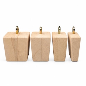 May include: Four light-colored wooden furniture legs arranged in a row. Each leg has a tapered, rectangular shape with a visible wood grain pattern and a small, metallic, circular attachment point fixed to the center of the flat top surface.