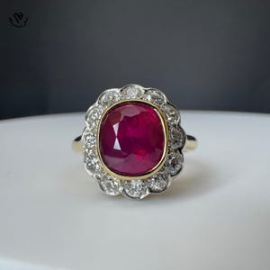 May include: A gold ring with a large, oval ruby centre stone, encircled by a halo of small, round diamonds. The ring's design is classic and elegant, suitable for special events. The ruby is a deep, rich red.
