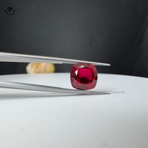 May include: A cushion-cut ruby, held by tweezers, showcasing a rich red colour. The gemstone's facets are clearly visible. The background is a soft white surface with a blurred, dark backdrop. The text "Naturesartdiamond" is visible.