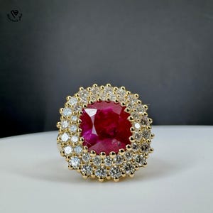 Può includere: Un anello d'oro con un grande rubino rosso scuro rotondo al centro, circondato da due aloni di diamanti più piccoli e scintillanti. L'incastonatura dell'anello è anch'essa in oro, creando un gioiello lussuoso e accattivante.