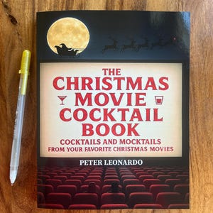 May include: A book titled "The Christmas Movie Cocktail Book" with a full moon and Santa's sleigh on the cover. The book's title is in red, with a pen to the left. The book cover features a theater scene.