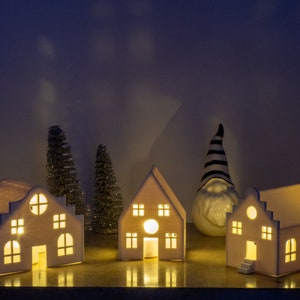 Könnte beinhalten: Ein Set aus vier weißen Keramikhäusern mit leuchtenden Fenstern, zwei kleinen Kiefern und einem weißen Keramik-Zwerg mit einem schwarz-weiß gestreiften Hut. Die Häuser sind von innen beleuchtet und erzeugen ein warmes und einladendes Leuchten.