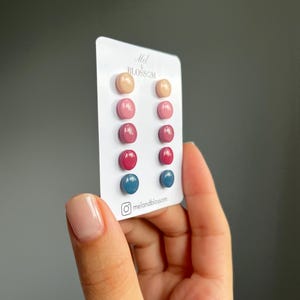 Pode incluir: Um cartão de brincos de botão em várias cores, incluindo pêssego, rosa, vermelho e azul. Os brincos são redondos e têm um acabamento brilhante. O cartão tem o texto "Mel & Blossom" e um nome de usuário do Instagram. Os brincos são segurados por uma pessoa.