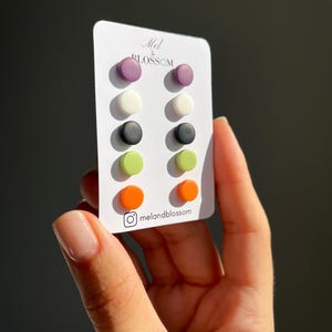 Op de afbeelding: Een set kleurrijke oorstekers in verschillende kleuren, waaronder paars, wit, zwart, groen en oranje. De oorbellen worden gepresenteerd op een witte kaart met de tekst "Mel & Blossom" en de Instagram-handle "melandblossom".