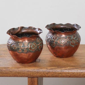 May include: Two rounded copper pots featuring a ruffled top edge and a decorative embossed floral vine pattern around the centre. The metal displays a warm reddish-brown tone with green oxidation accents within the recessed floral details.