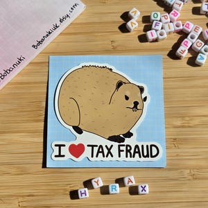 Puede incluir: Una pegatina con un hiracoide de dibujos animados con el texto "I ❤️ TAX FRAUD" sobre un fondo azul claro. El hiracoide es marrón con un contorno blanco. La pegatina está sobre una superficie de madera.