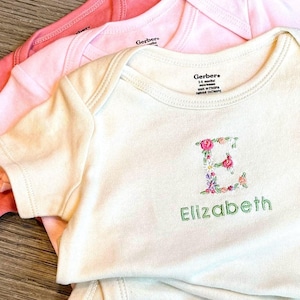 May include: A white onesie with a floral letter E and the name Elizabeth embroidered on the front. The onesie is folded on top of two other onesies, one pink and one coral.