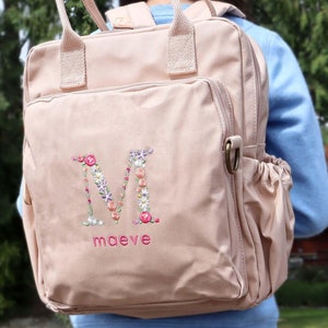 May include: A light pink canvas backpack with a floral embroidered letter "M" and the name "maeve" embroidered below.