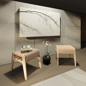 May include: Two light wood slatted stools with a simple design. The stools are in a modern living room setting with a large framed print of a tree branch on the wall behind them.