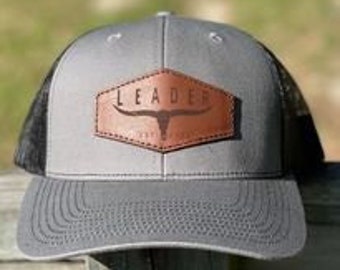Könnte beinhalten: Eine graue und schwarze Trucker-Kappe mit einem braunen Leder-Patch, auf dem "LEADER" mit einem Stierkopf-Design steht.