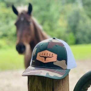 Può includere: Un cappello da camionista mimetico con una toppa in pelle marrone che dice "LEADER" in lettere bianche. La toppa presenta anche una testa di toro stilizzata.