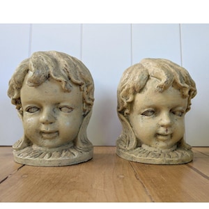 Könnte beinhalten: Zwei antike, cremefarbene Cherubinkopfskulpturen. Jede hat detaillierte Gesichtszüge, einschließlich geschlossener Augen und eines leichten Lächelns, mit lockigem Haar und einem gerüschten Kragen. Die Skulpturen werden auf einer Holzoberfläche ausgestellt.