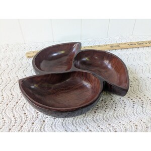 May include: A dark brown, three-section wooden serving dish. The dish has a clover-like shape with rounded compartments. The wood grain is visible, adding texture to the piece. The dish is likely used for serving snacks or appetizers.