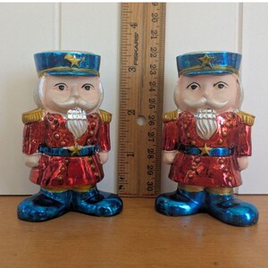 Pode incluir: Duas figuras vintage de quebra-nozes de Natal. Cada figura é pintada com um casaco vermelho, botas e chapéu azuis e um bigode branco. Os chapéus têm uma estrela dourada. Uma régua é colocada entre as figuras.