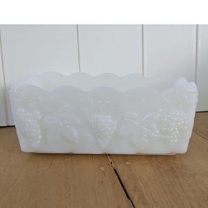 Puede incluir: Un plato rectangular de vidrio blanco con borde festoneado. Los lados están decorados con un diseño de uvas y hojas. El plato está sobre una superficie de madera.