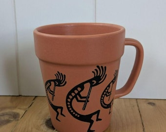 Puede incluir: Una taza de cerámica color terracota con forma de maceta, que presenta un borde ancho y un asa lateral. El exterior está decorado con tres siluetas gráficas negras de una figura tocando la flauta en un patrón repetido alrededor.